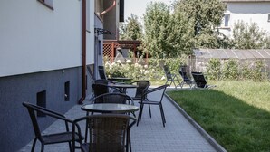 Un patio al aire libre con sillas y mesas de mimbre, un pequeño jardín y un edificio al fondo.