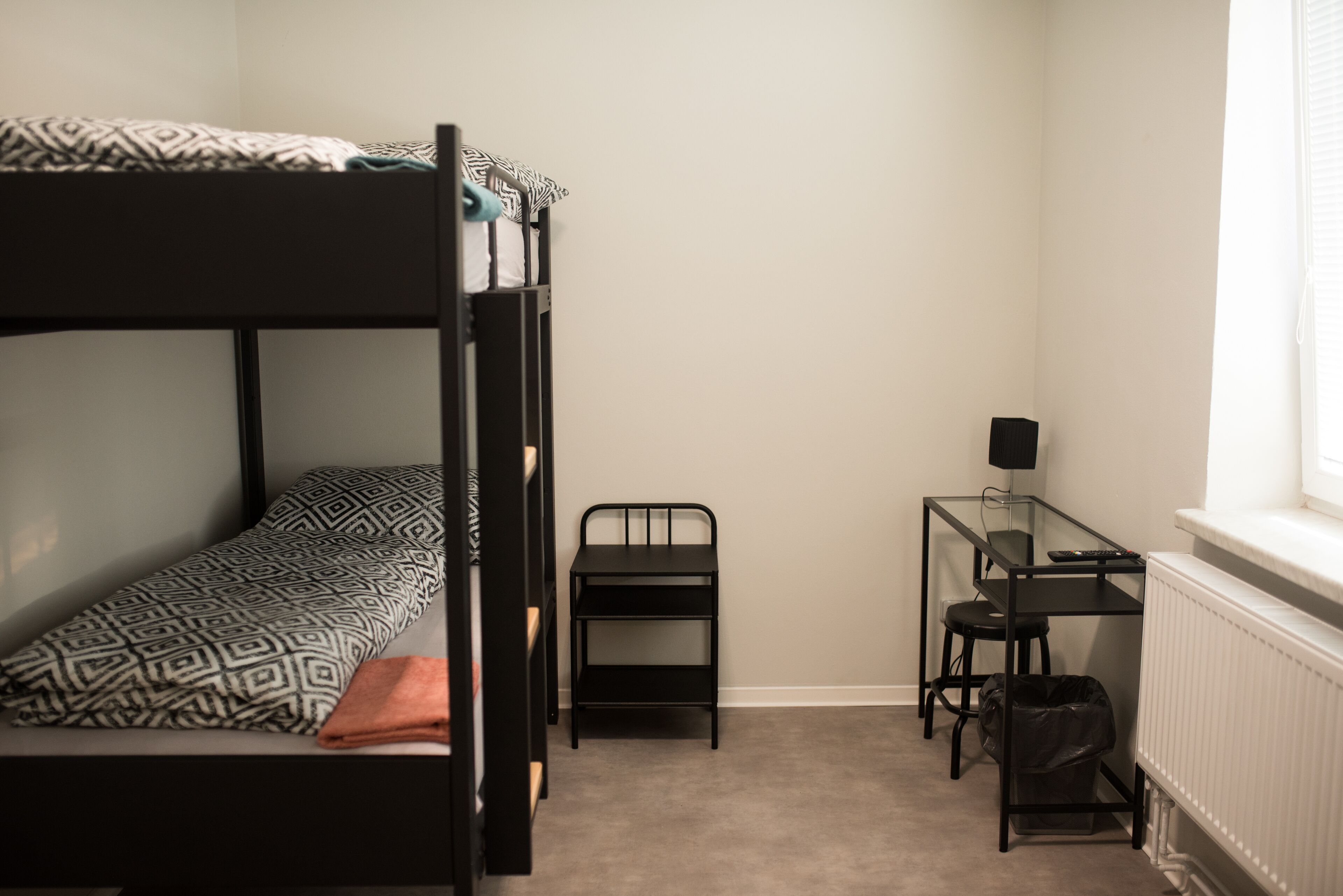A dormitory room with a bunk bed, a desk, a chair, and a radiator.