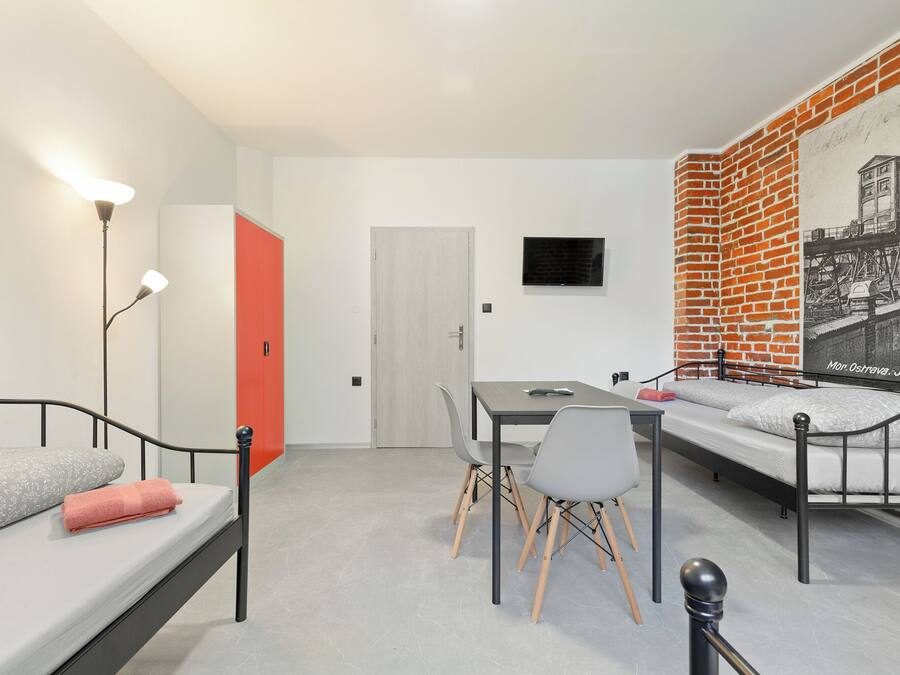 A modern room with a bed, a desk, and a chair. There is a door, a wall-mounted TV, and a brick-patterned wall.
