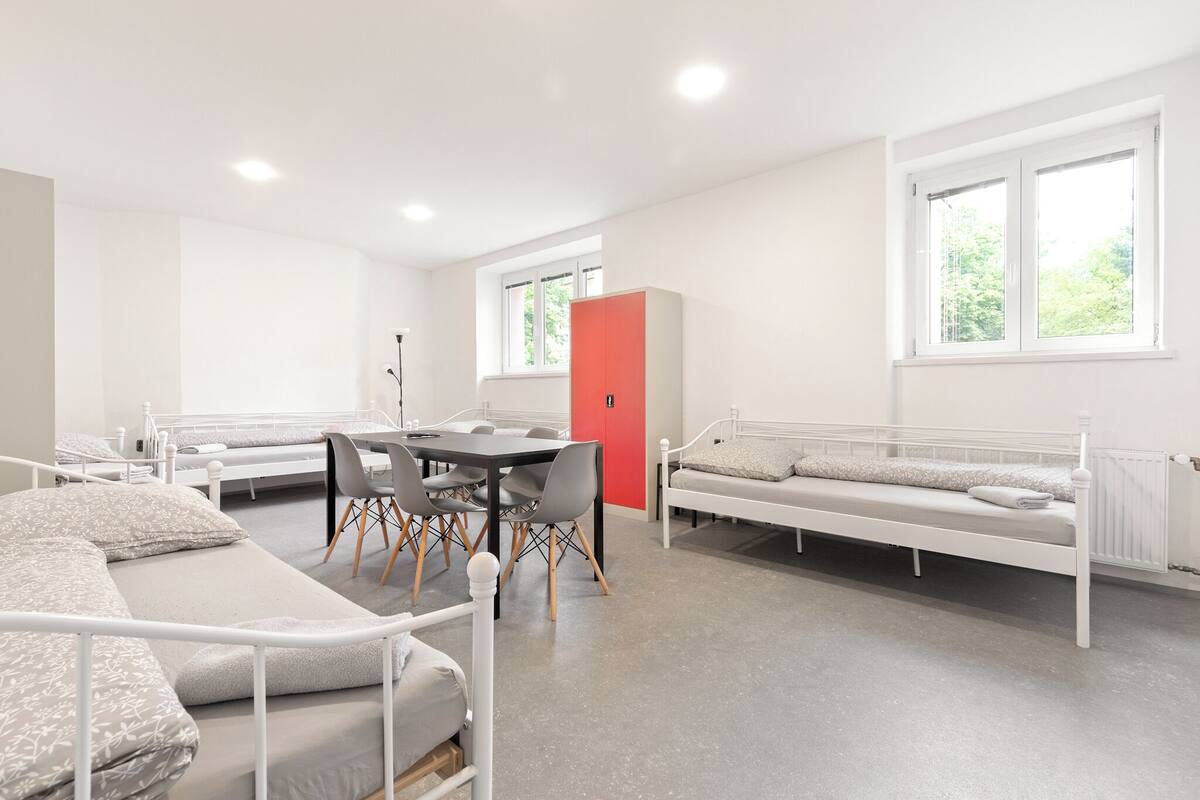 A modern, well-lit room with a dining table, chairs, and a sofa. There are beds with pillows and a red cabinet.