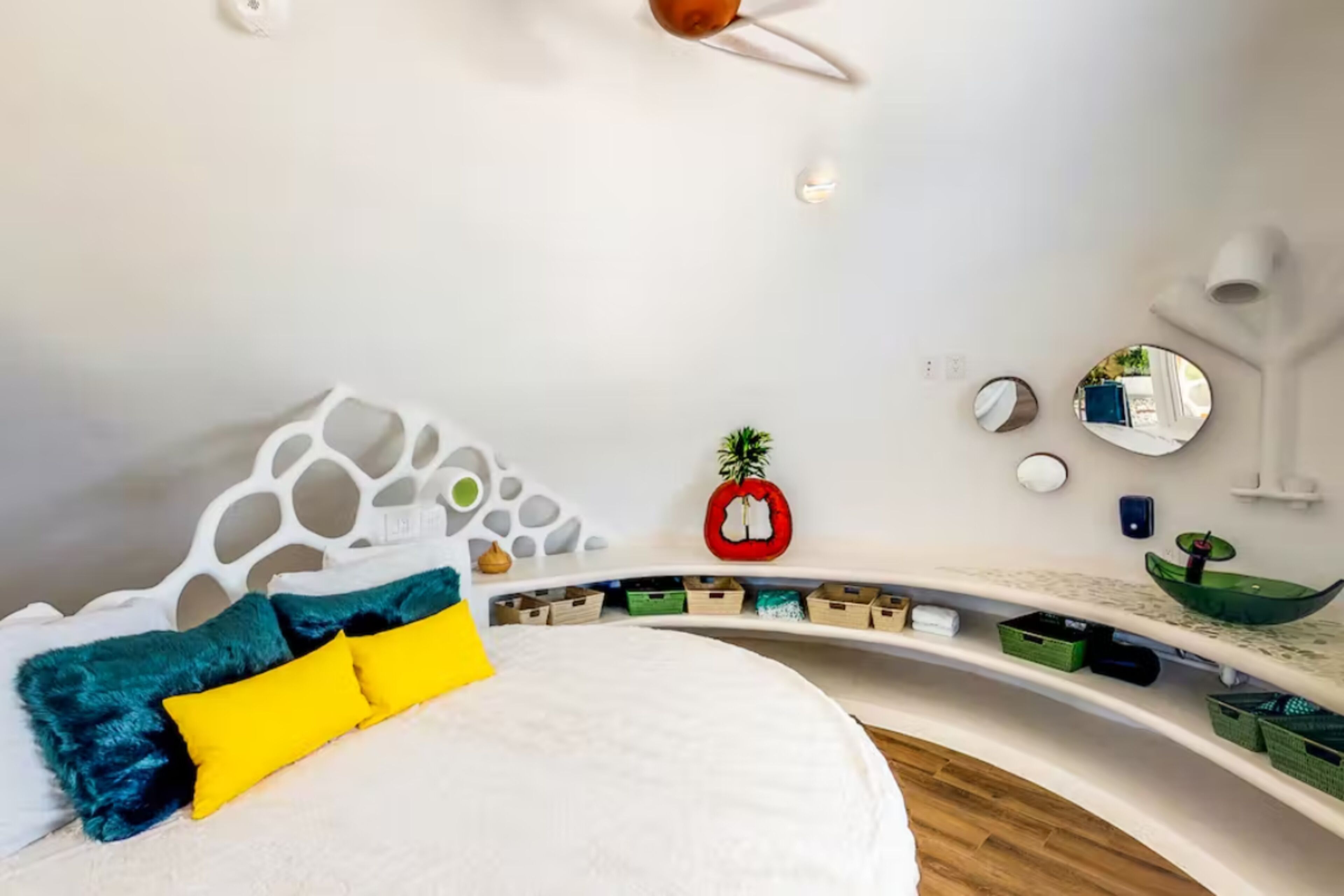 A modern bedroom with a white bed, colorful pillows, a unique white headboard, and a curved shelving unit with storage boxes.