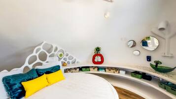 A modern bedroom with a white bed, colorful pillows, a unique white headboard, and a curved shelving unit with storage boxes.