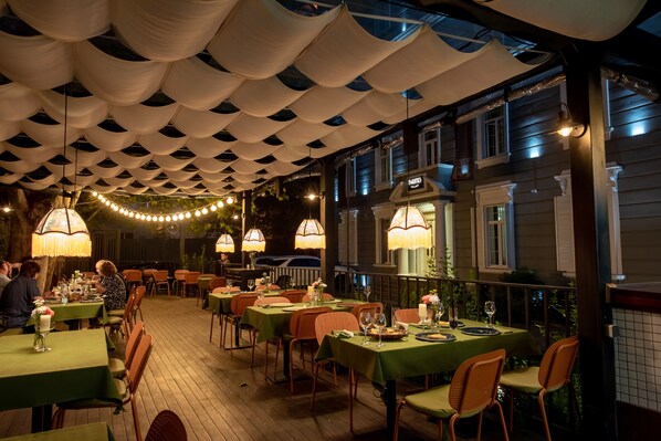 Un espace de restauration en plein air, avec des tables et des chaises, des guirlandes lumineuses et une partie abritée offrant une vue sur un bâtiment.