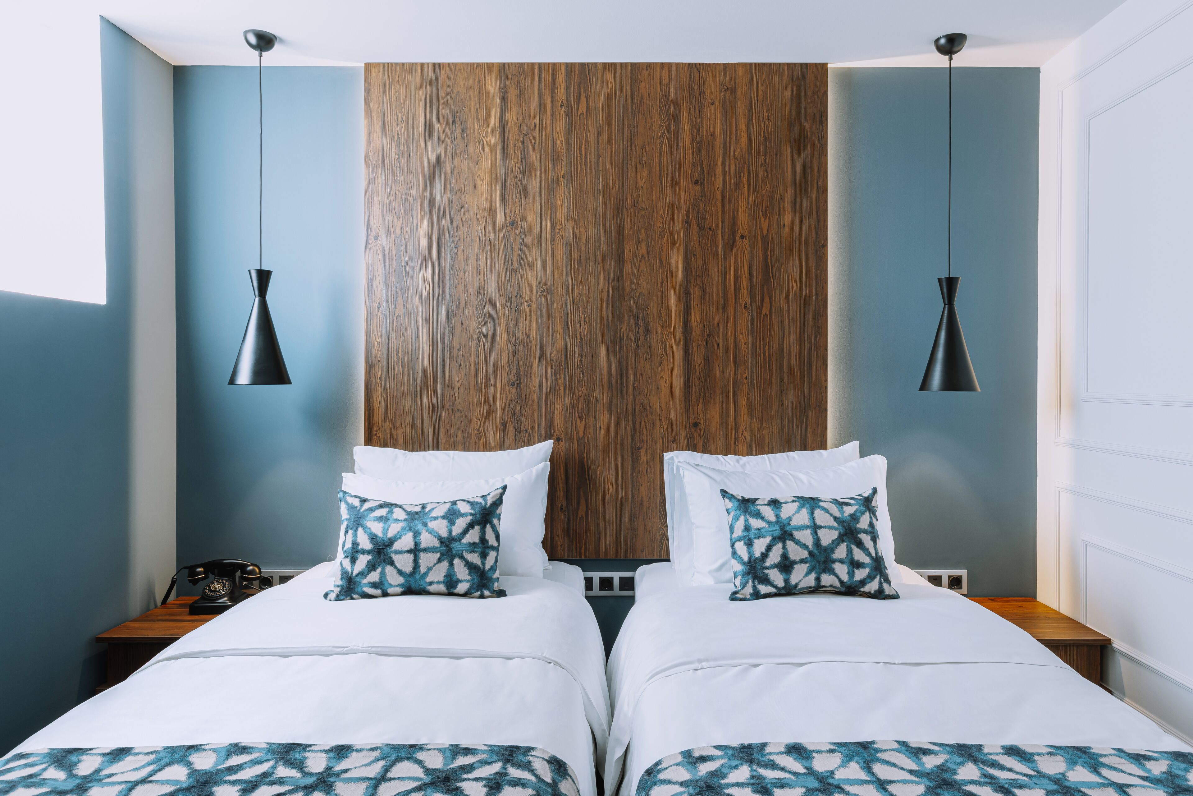 Un lit double avec du linge de lit blanc et des coussins à motifs bleus, une tête de lit en bois et deux lampes suspendues noires.