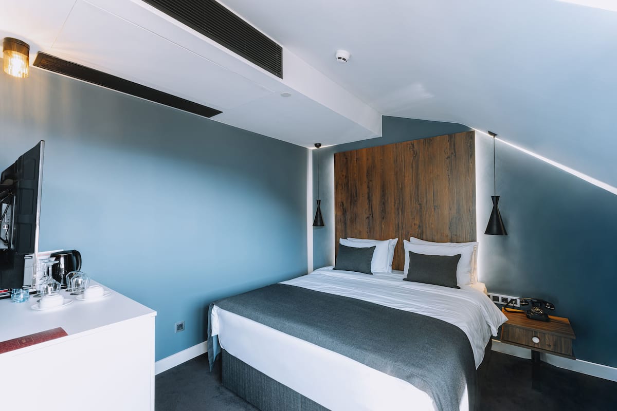 A modern hotel room with a large bed, a wooden accent wall, a bedside table with a lamp, and a desk with a coffee maker.