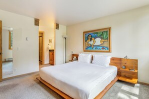 Una camera d'hotel con un letto grande, una testiera in legno, comodini, un tavolino da notte, un quadro appeso al muro e una porta che dà su un'altra stanza.