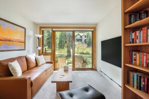 A modern living room with a brown leather sofa, a wooden coffee table, a black ottoman, a bookshelf, and a large window with a view of greenery.