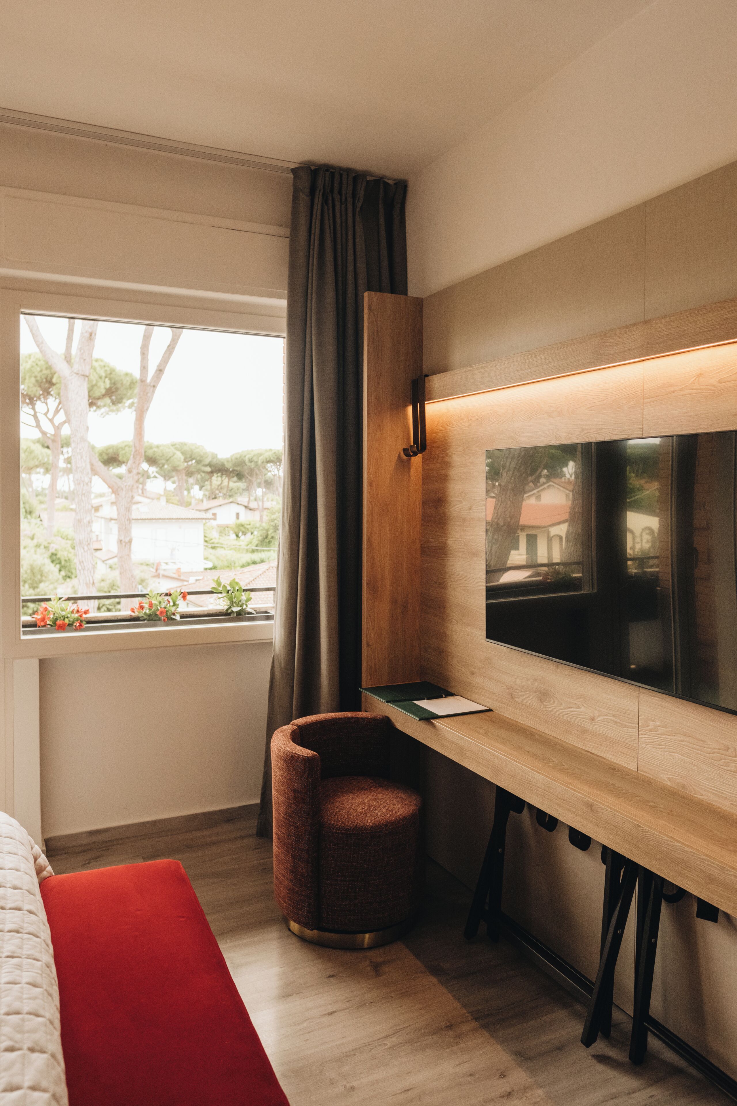 Una habitación moderna con una cama, un banco rojo, un escritorio de madera y un ventanal con cortinas.
