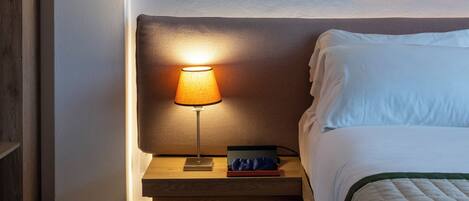 A neatly made bed with a white comforter, a bedside lamp, and a pair of slippers on the floor.