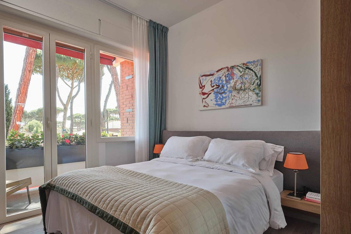 A hotel room with a bed, a nightstand, a lamp, a painting, and a view of the outside.