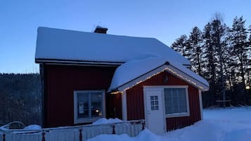 A red wooden cabin with a snow-covered roof, a white door, and a small porch area.