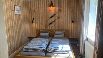 Una habitación con pared de listones de madera, una cama con ropa de cama a rayas, dos mesitas de noche y una lámpara colgante.