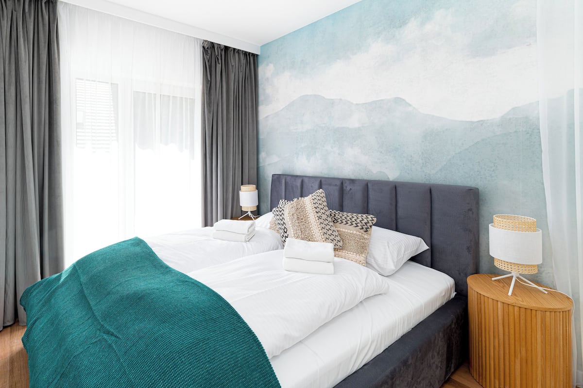 Een net opgemaakt bed met witte lakens, een blauwgroene plaid en decoratieve kussens. Twee nachtkastjes met lampen en een wandlamp. De kamer heeft een groot raam met transparante gordijnen en een berglandschap op de muur.