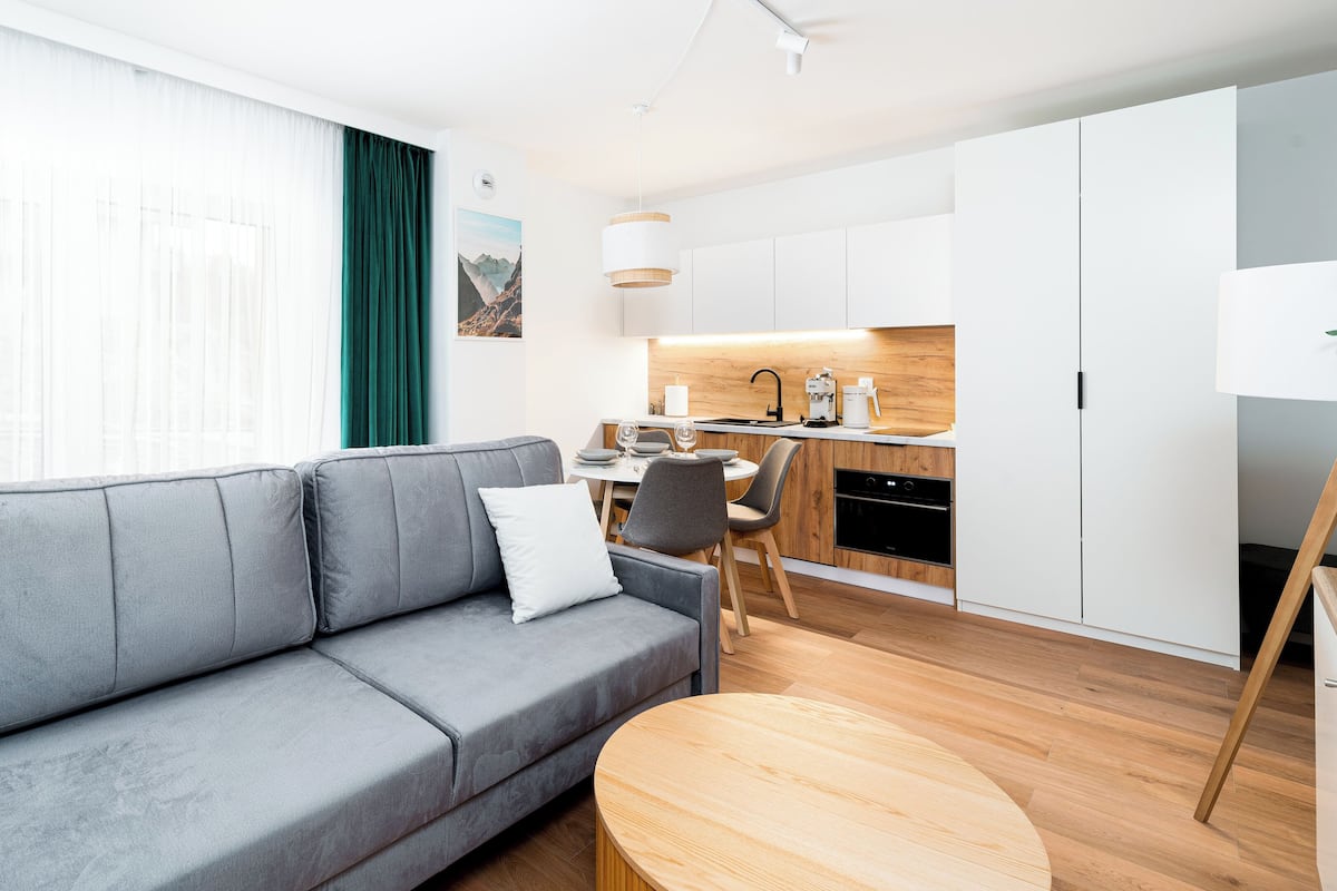 Een moderne woonkamer met een grijze bank, een klein houten tafeltje en een keukenhoek met witte kastjes en een houten aanrecht.