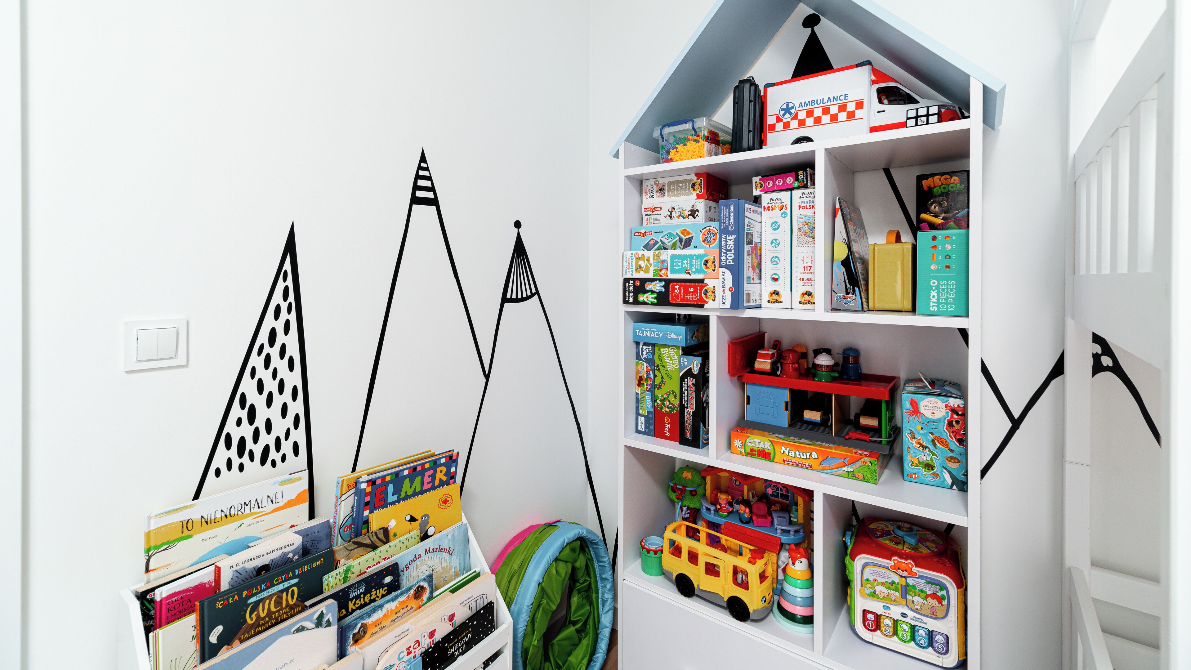 Ein Kinderzimmer mit weißer Wand, einem Bücherregal voller Spielzeug und Büchern sowie einer zeltartigen Konstruktion.
