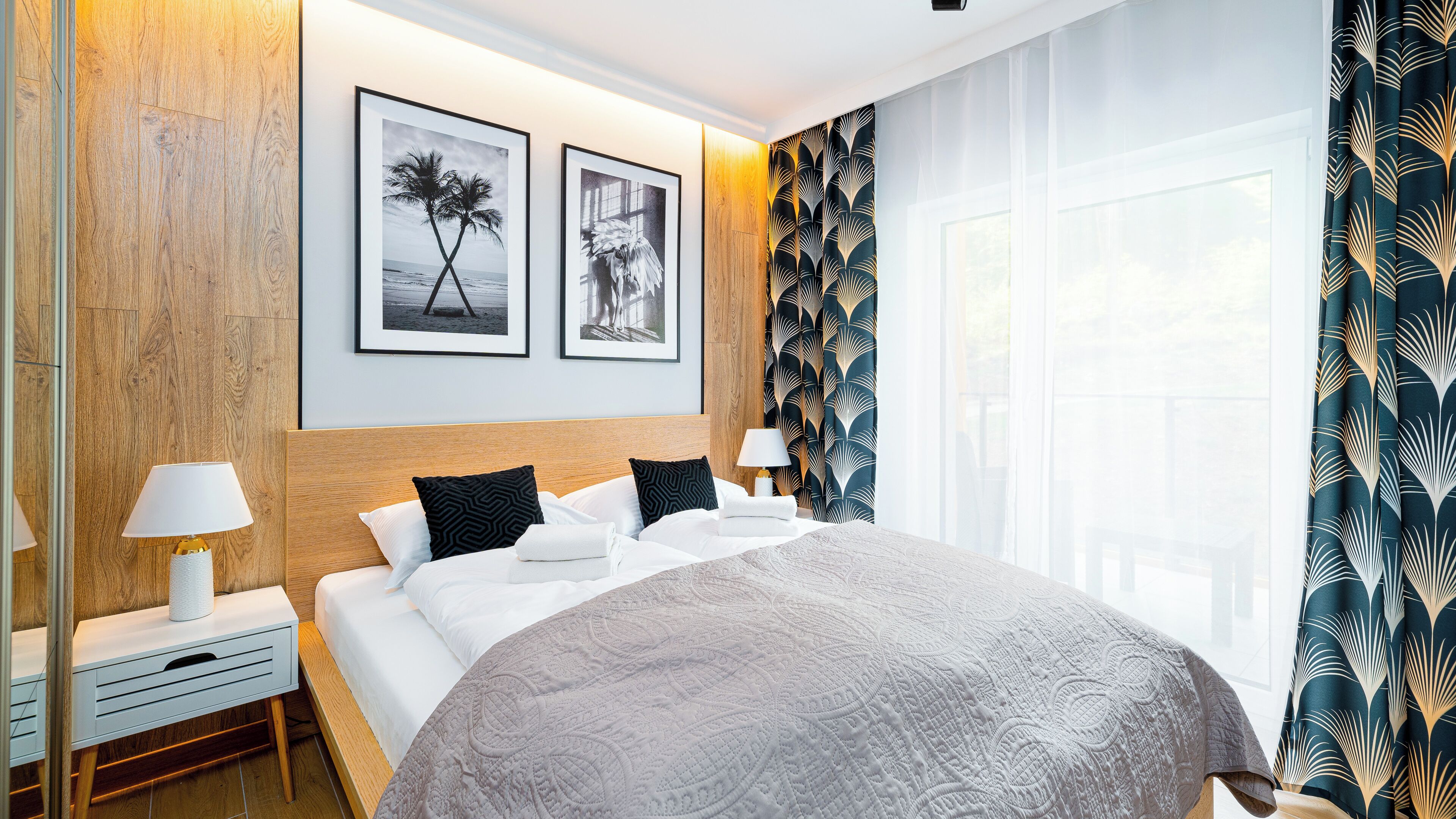 A hotel room with a large bed, two framed pictures on the wall, a nightstand with a lamp, and a window with curtains.