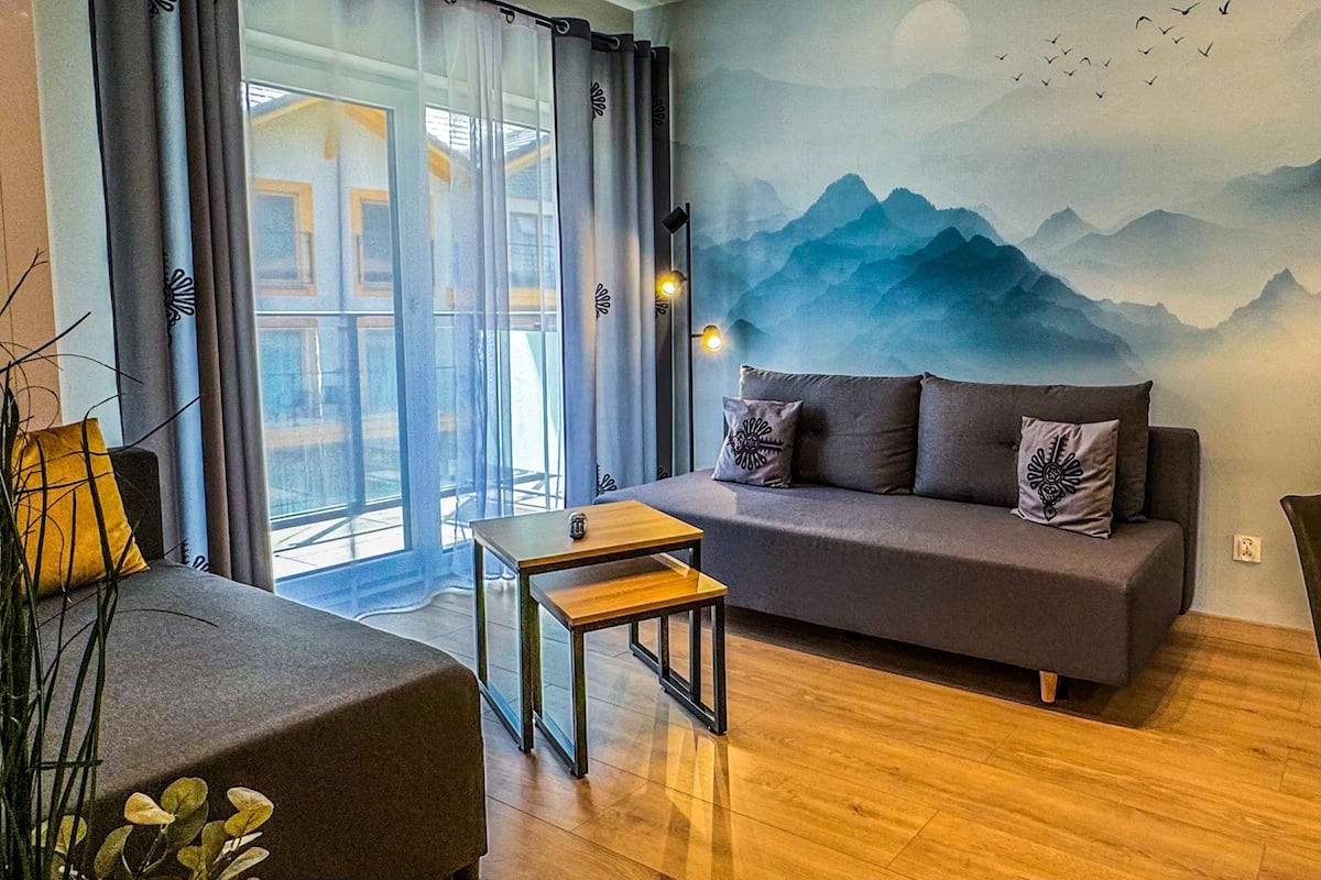 Een moderne woonkamer met een grijze bank, een klein houten tafeltje en uitzicht op bergen door een groot raam.