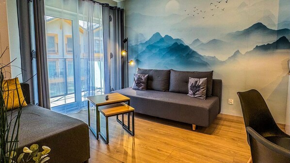 Una sala moderna con un sofá gris, una pequeña mesa de madera y vistas a montañas a través de un ventanal grande.