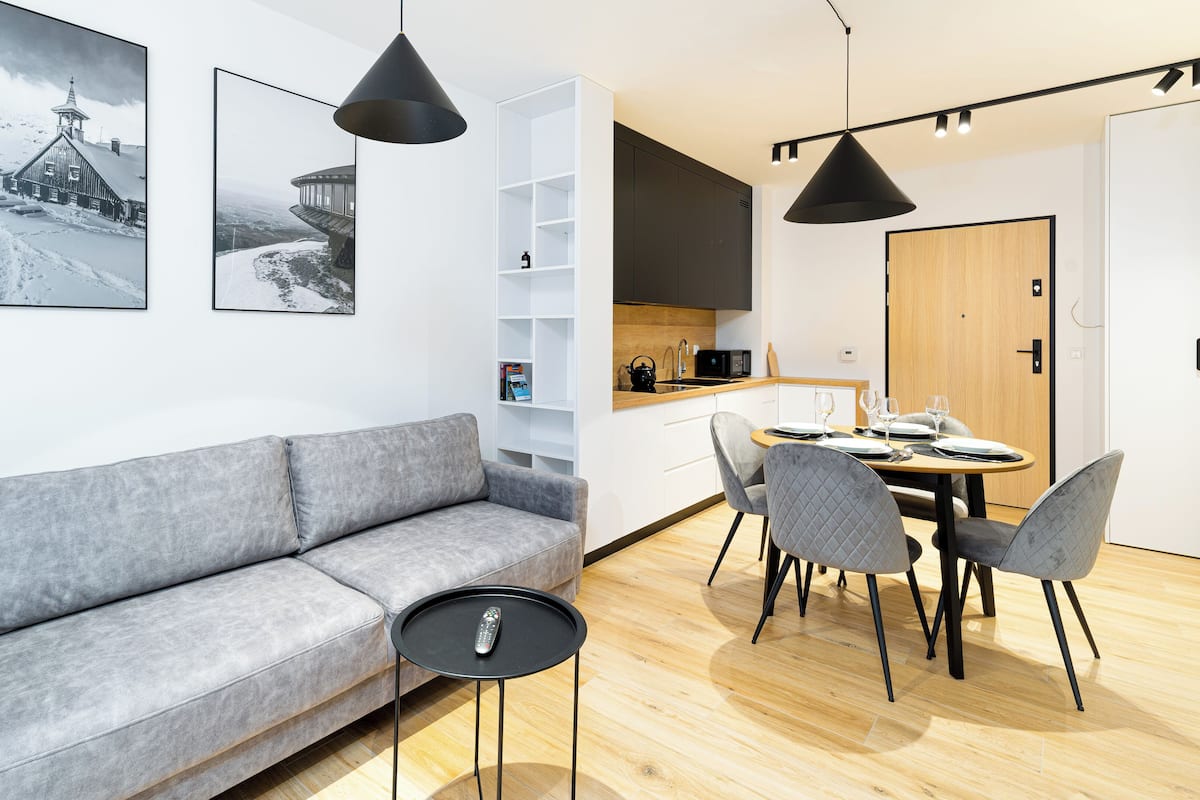 Een moderne woonkamer met een grijze bank, een klein zwart bijzettafeltje en een eethoek met een ronde tafel en grijze stoelen. Boven de bank en eettafel hangen twee hanglampen.