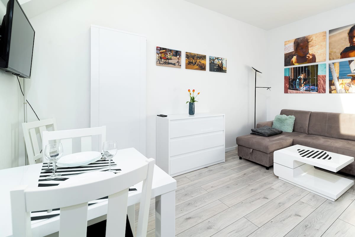 Een moderne woonkamer met een witte eettafel, een bank en een salontafel. Er hangen drie ingelijste foto's aan de muur en een tv is aan de muur bevestigd.