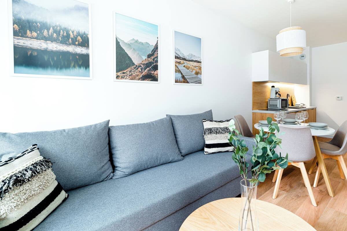 Een moderne woonkamer met een grijze bank, een eettafel met stoelen, en een keuken met een magnetron en koffiezetapparaat.