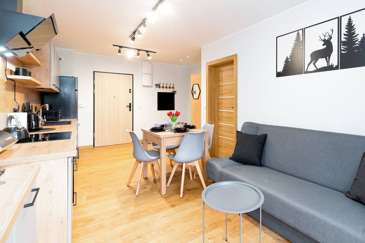 Een moderne woonkamer met een grijze bank, een klein rond tafeltje en een eetgedeelte met een houten tafel en stoelen.