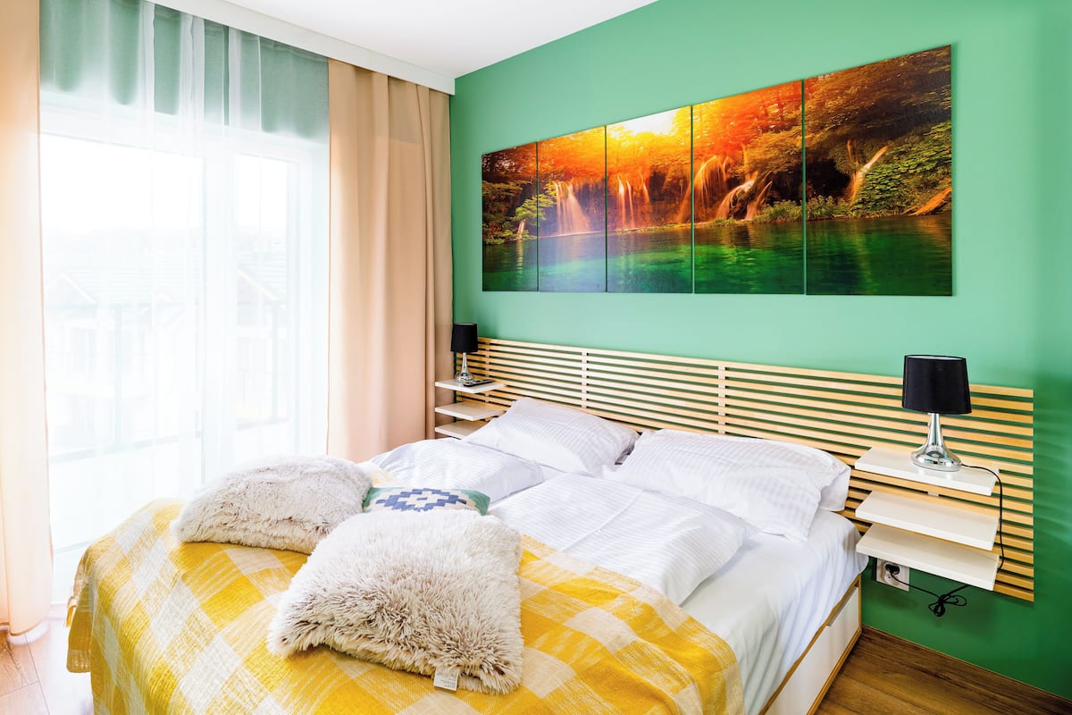 Een keurig opgemaakt bed met een geruit sprei en twee zachte kussens, een nachtkastje met een lamp, en een groot schilderij van een waterval aan de muur.