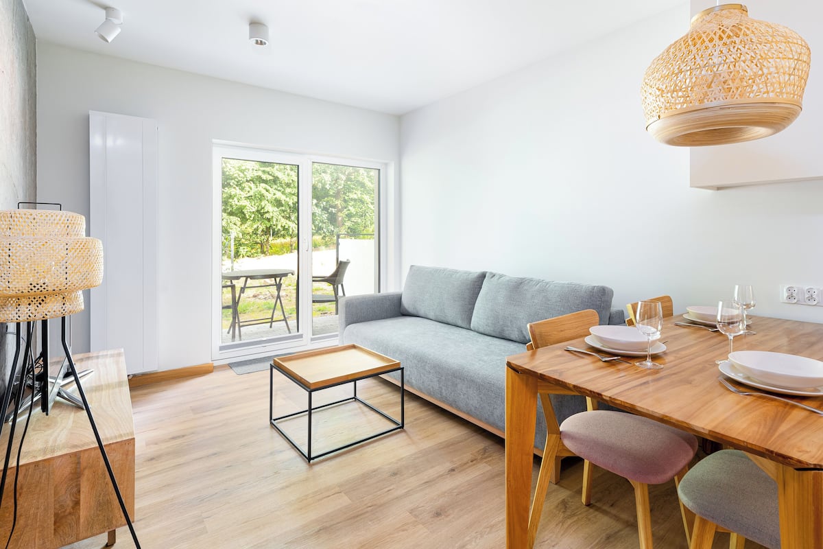 Een moderne leefruimte met een houten eettafel, een grijze bank en een glazen deur naar een buitenruimte.