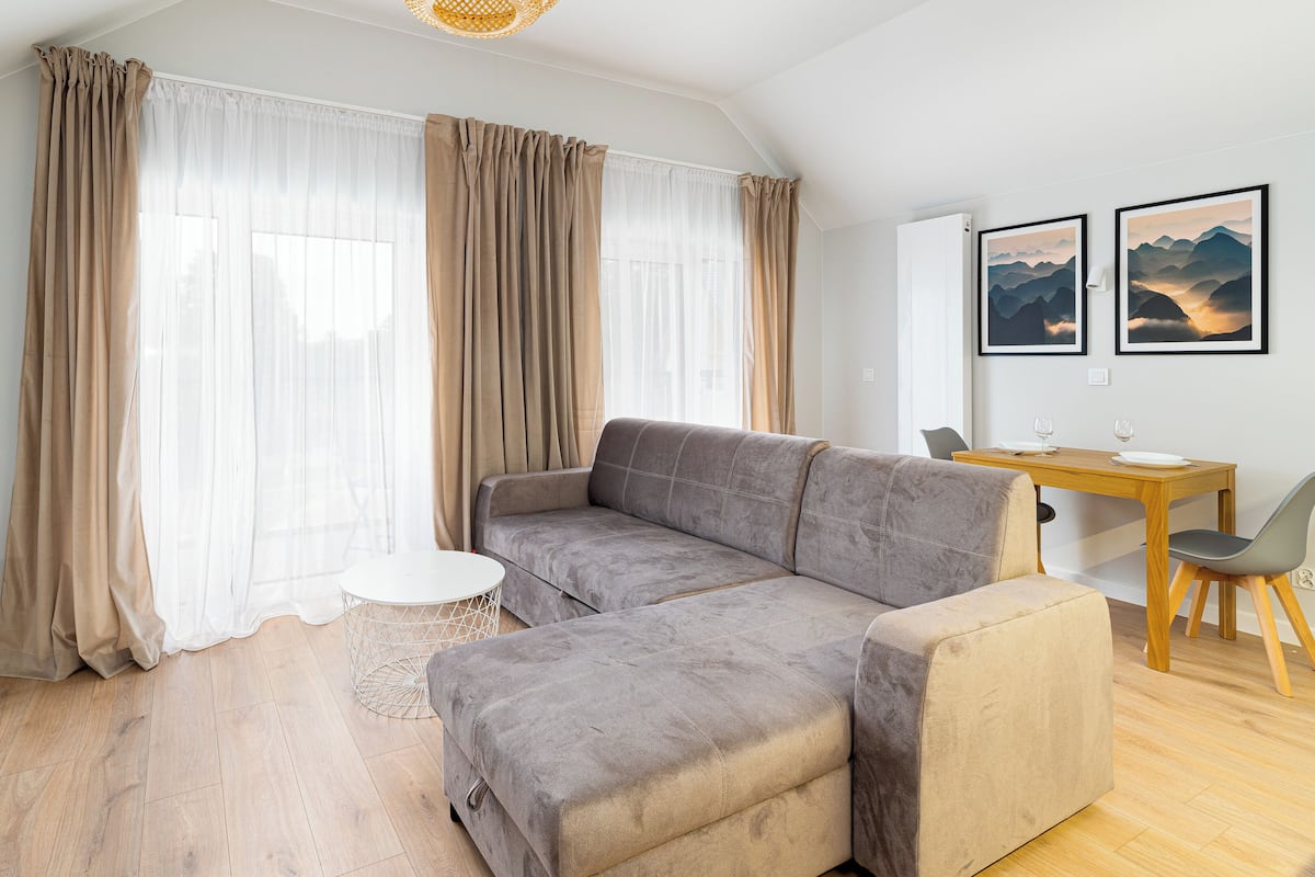 Een moderne woonkamer met een grijze bank, een kleine ronde tafel en een eethoek met een houten tafel en stoelen.
