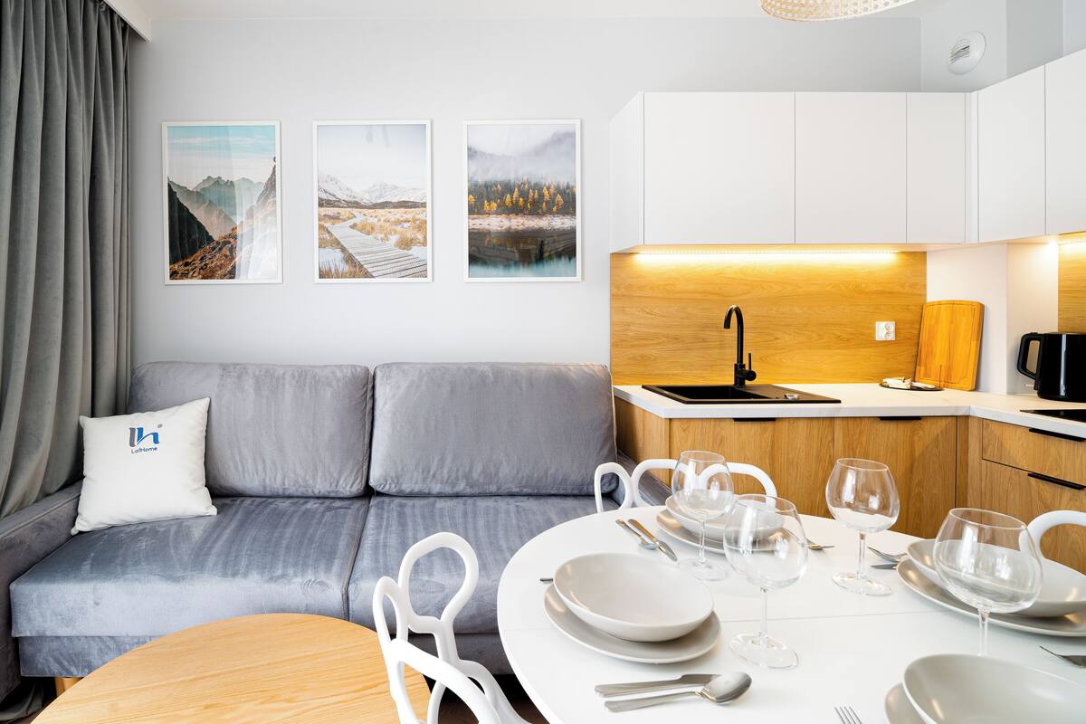 Een moderne woonkamer met een grijze bank, een eettafel met borden en glazen, en een keuken met houten kasten en een zwarte waterkoker.