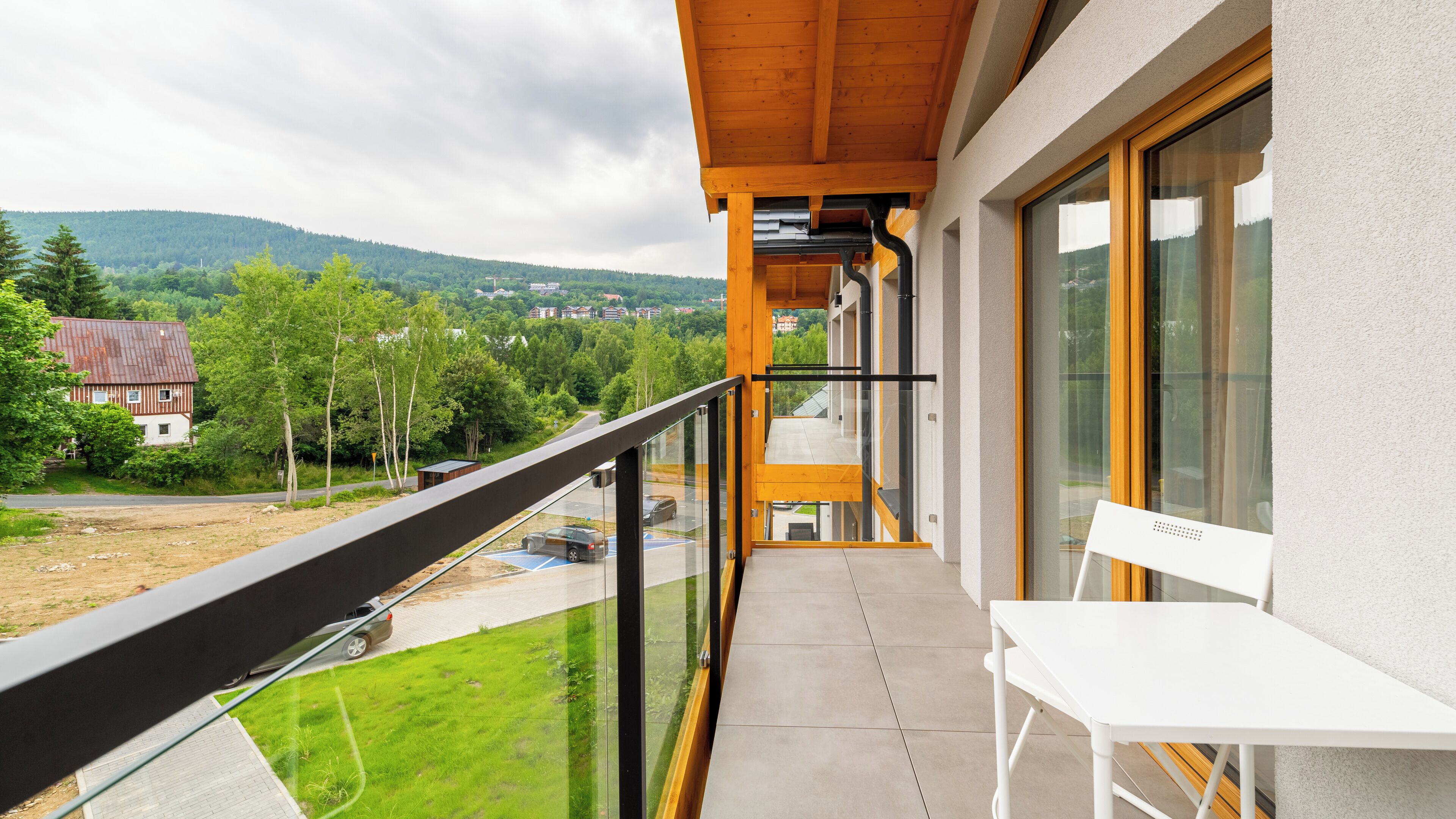 Un balcon avec une chaise blanche, une balustrade en verre et une vue sur un paysage verdoyant, avec des arbres et un bâtiment situé au loin.