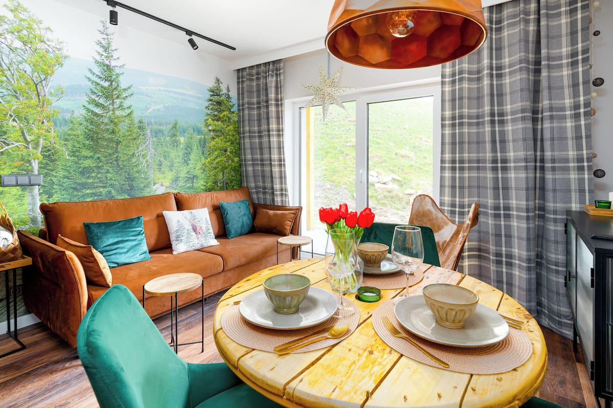 Een gezellige woonkamer met een bank, een eettafel gedekt met borden en een vaas rode bloemen, en een groot raam met uitzicht op een bos.