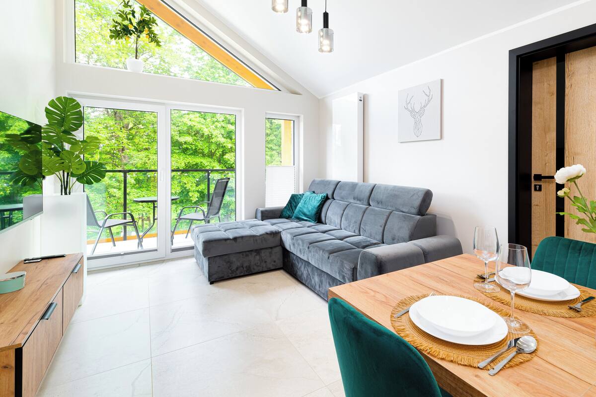 Een moderne woonkamer met een grijze bank, een houten eettafel gedekt voor twee personen en een groot raam met uitzicht op een groene tuin.