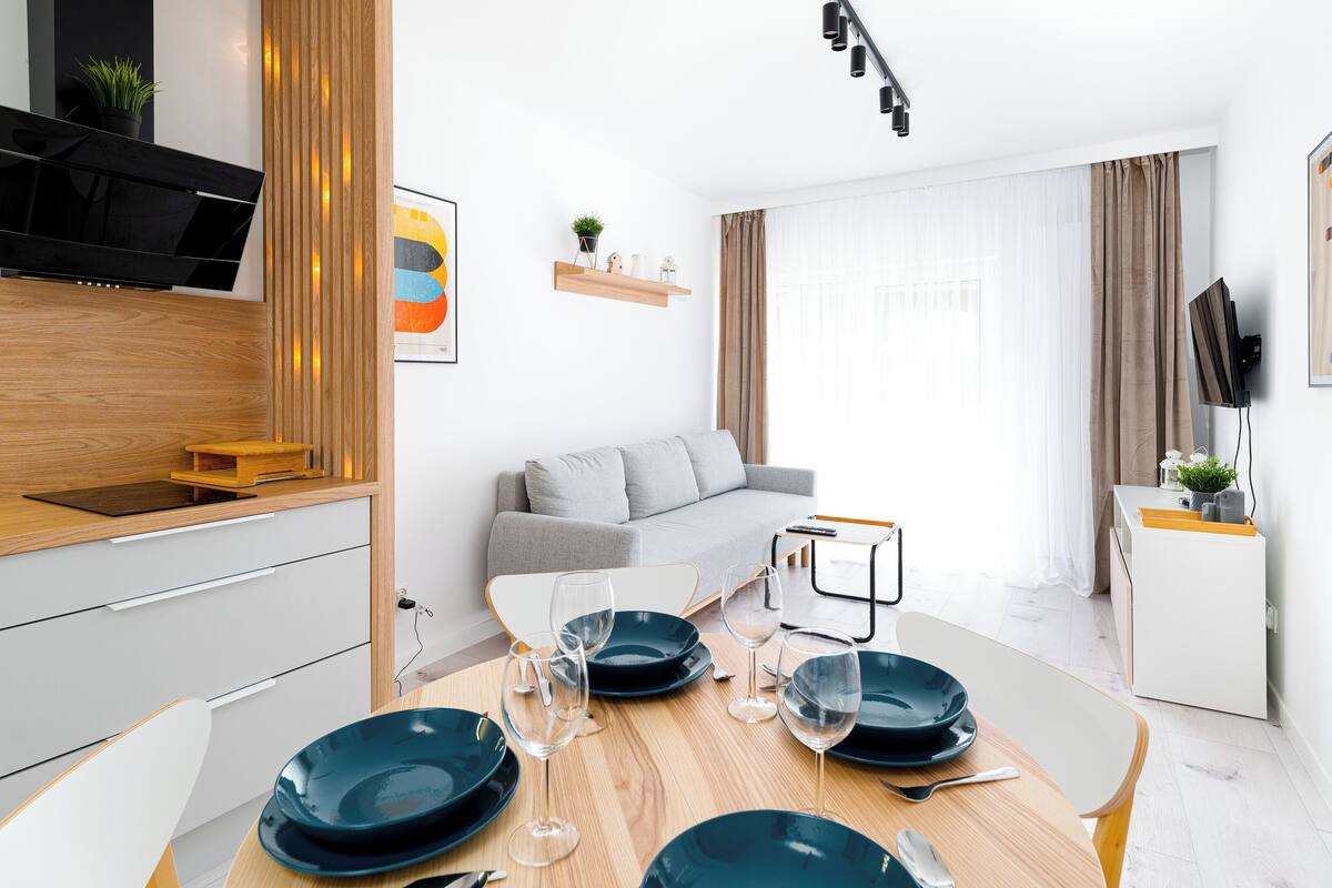 Een moderne keuken met een ingebouwde kookplaat, een eettafel gedekt met borden en glazen, een grijze bank en een aan de muur bevestigde televisie.