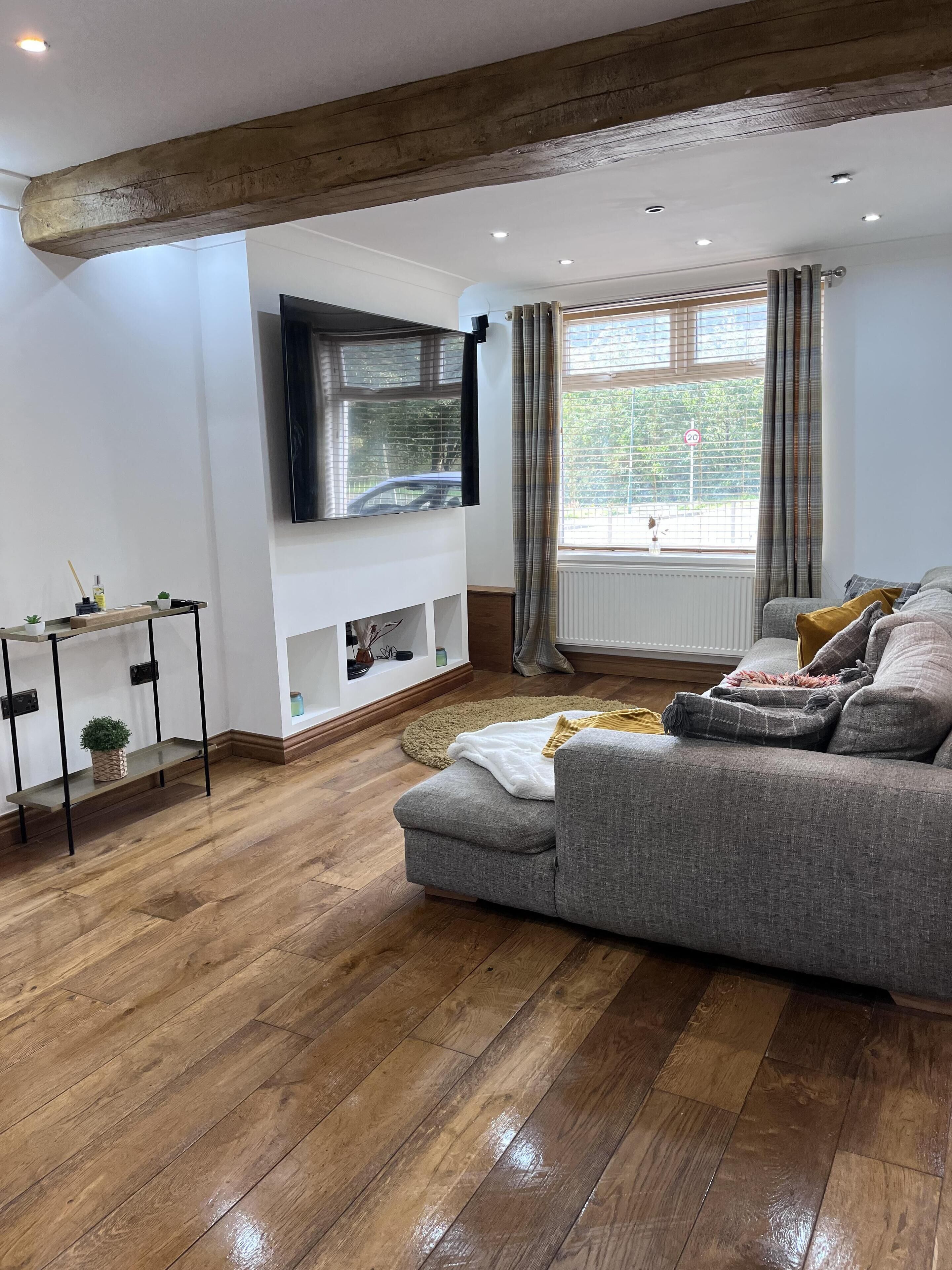 Un salon moderne avec un canapé gris, un plancher en bois, un téléviseur fixé au mur et une fenêtre avec des rideaux.