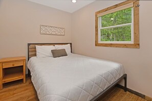 A bedroom with a bed, a nightstand, a window with a view of greenery, and a decorative wall piece. - Pet Friendly - Close to Town - w/ Deck & Fire Pit (Lake George)