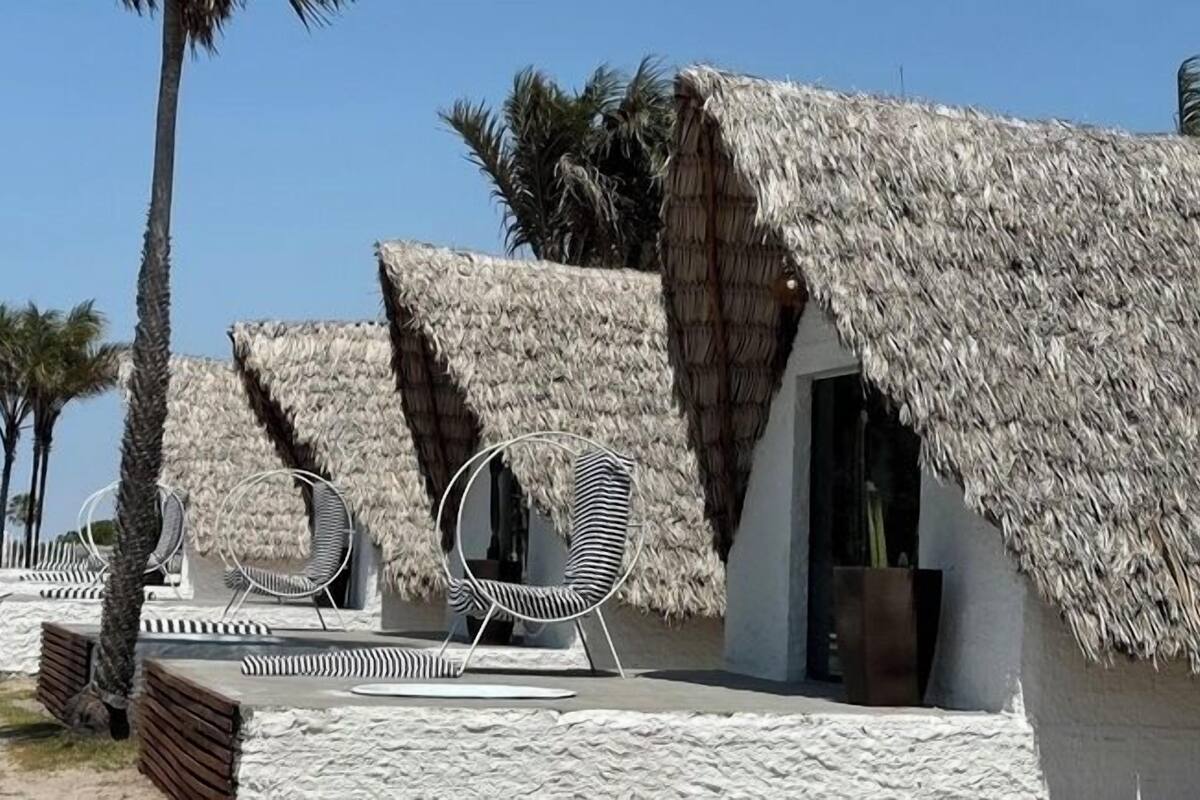 Cabañas con techo de paja, sillas de descanso y una pequeña mesa, rodeadas de palmeras.