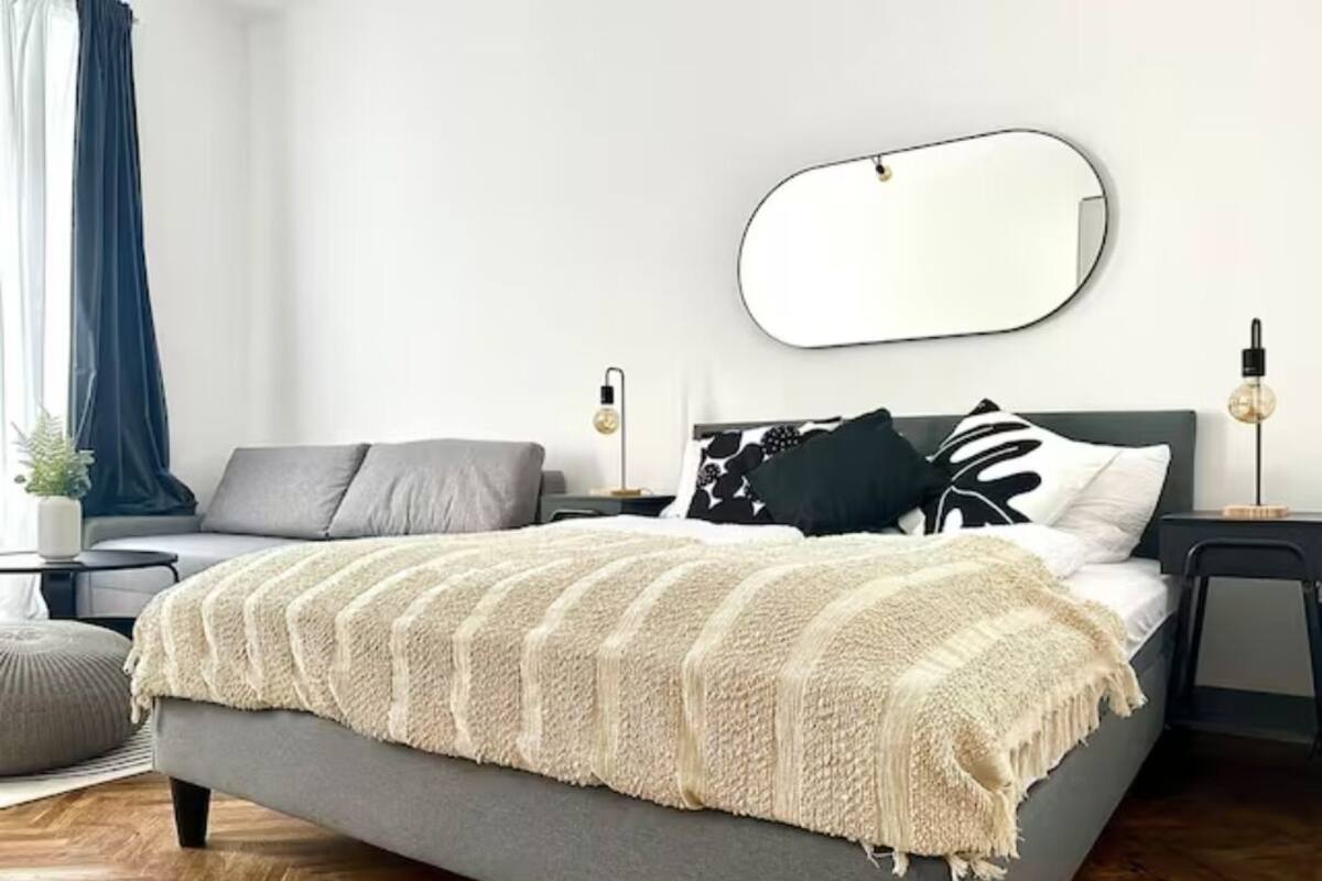 A modern bedroom with a grey bed, a large mirror, and a bedside table with a lamp.