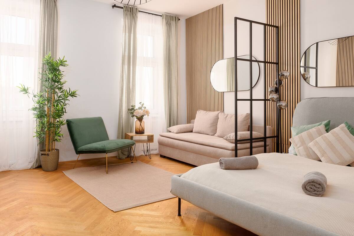 A modern bedroom with a wooden floor, a green chair, a sofa, a bed with pillows, a bedside table with a vase, and a mirror on the wall.