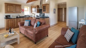 A spacious living room with a brown leather sofa, a wooden coffee table, and a kitchen area with wooden cabinets and a stainless steel refrigerator. - Smithy Cottage (Alnwick)