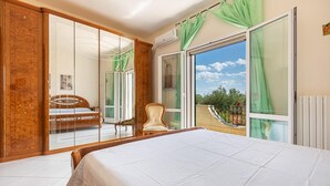 A bedroom with a large bed, a wooden wardrobe, a chair, and a balcony with a view of greenery. - 3634 Villa Dolce - App1 by Barbarhouse (Ostuni)