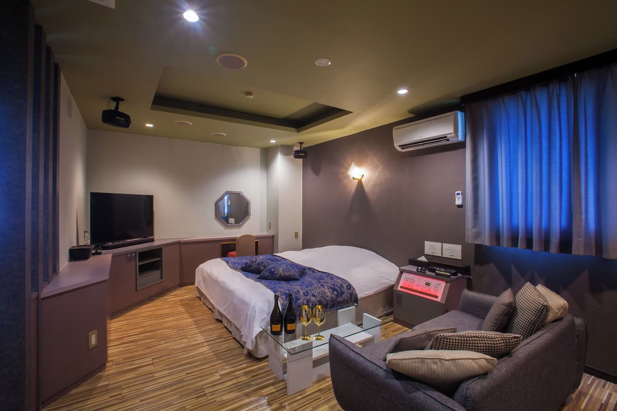 A modern hotel room with a large bed, a flat-screen TV, a desk with a computer, and a seating area with a couch and armchairs.