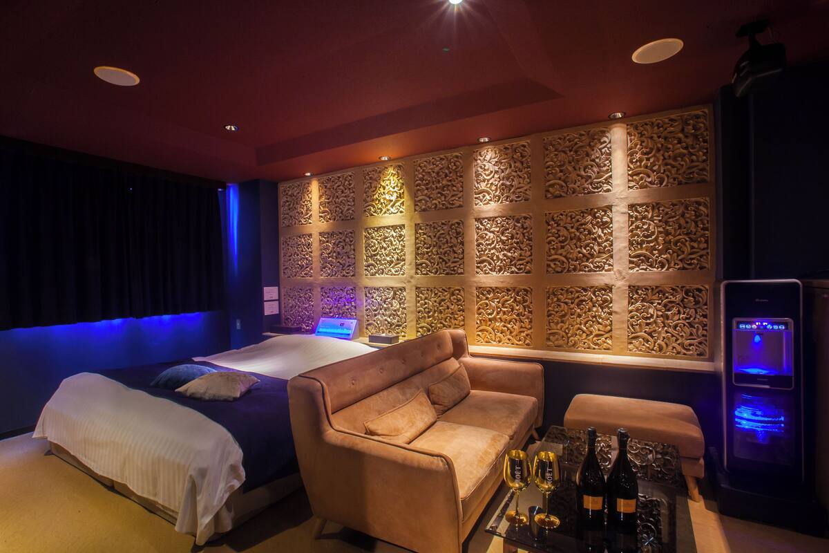 A room with a bed, a sofa, a table with bottles, and a wall with intricate carvings.