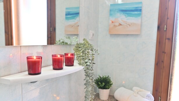 Un baño con encimera de mármol blanco, tres velas rojas, una planta en maceta y una toalla blanca enrollada.