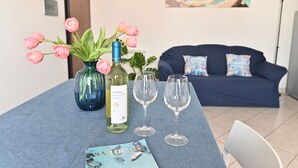 Una mesa de comedor con una botella de vino, dos copas y un menú. Un jarrón azul con flores rosas está sobre la mesa. En el fondo, un sofá azul oscuro con cojines estampados.