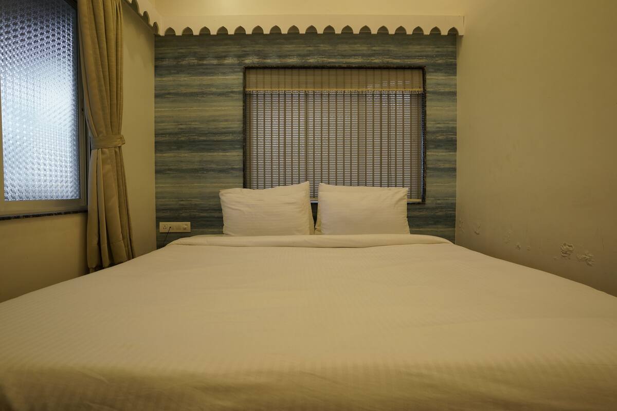 A neatly made bed with a headboard against a textured wall, a window with blinds, and a curtain on the left.