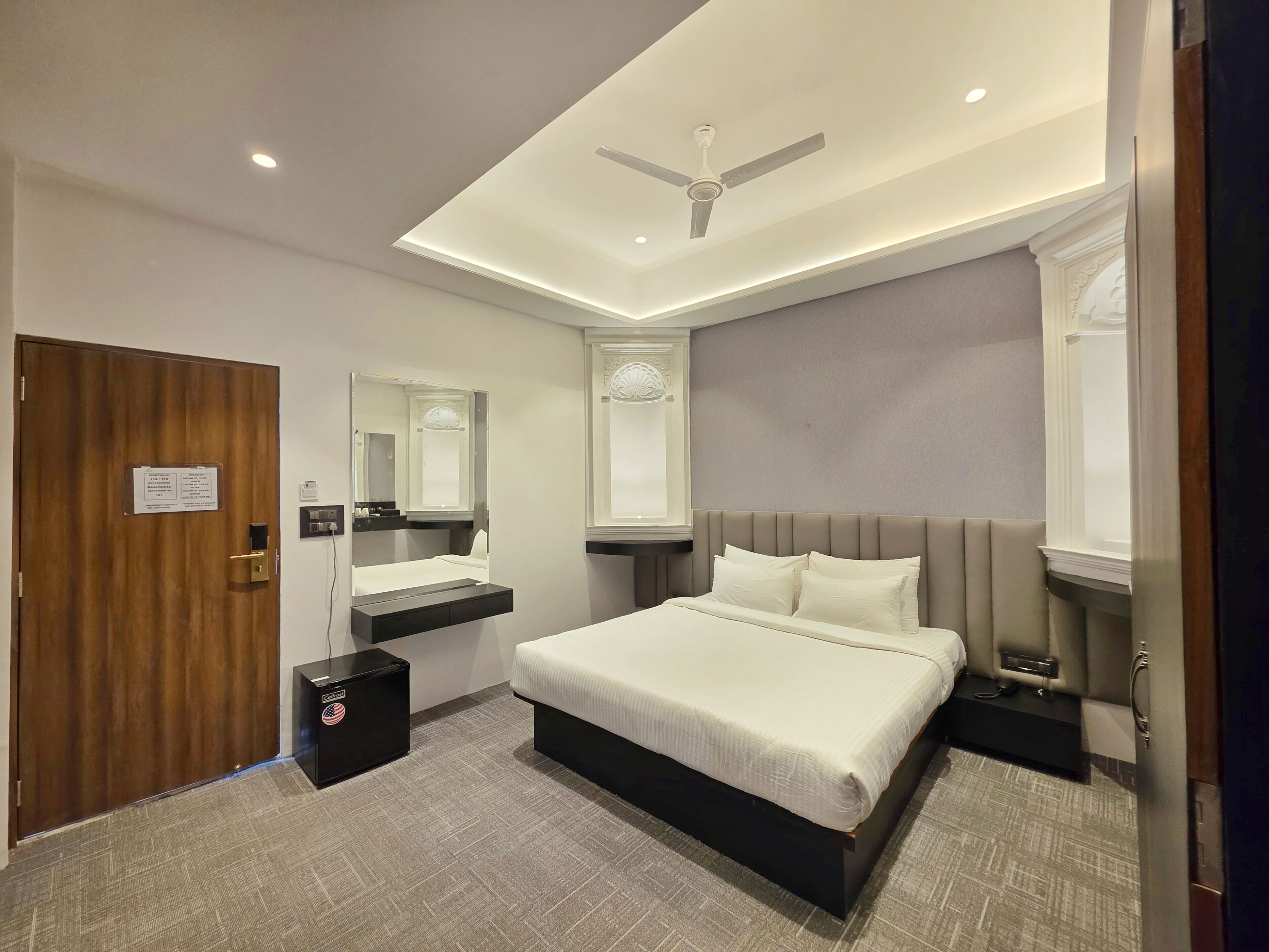 Une chambre d’hôtel avec un lit, des tables de chevet, un miroir, un ventilateur de plafond et une porte avec une pancarte.