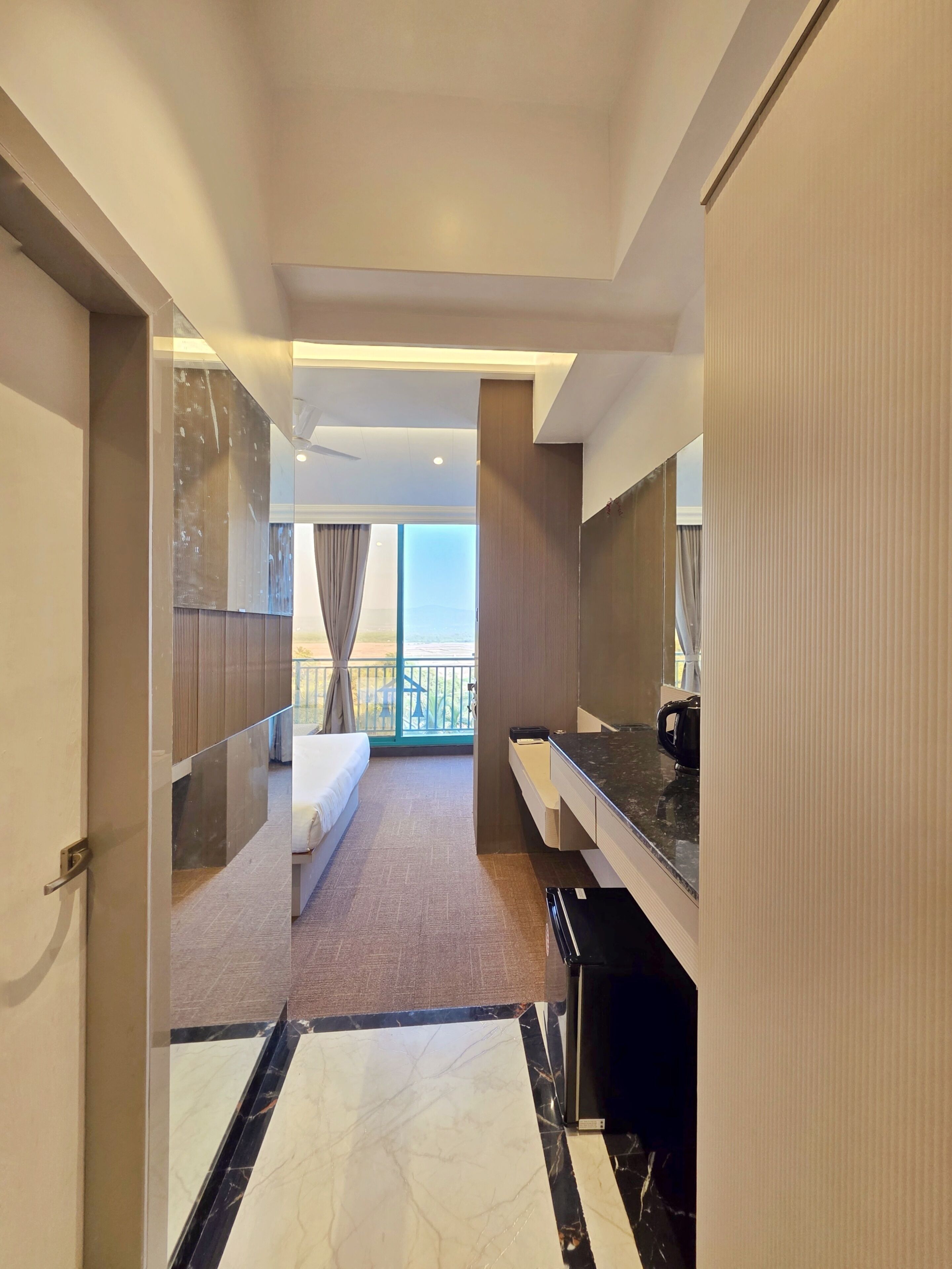 A hotel room with a bed, a bathroom with a mirror and sink, a balcony with curtains, and a view of the outdoors.