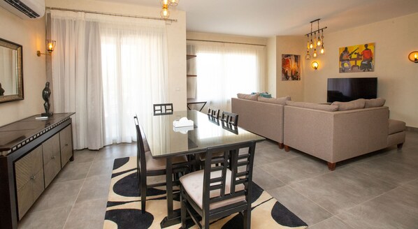 A modern living room with a glass dining table, beige sofa, and a flat-screen TV.