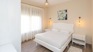 Een moderne slaapkamer met een groot bed, witte gordijnen, een schilderij en twee bedlampjes.